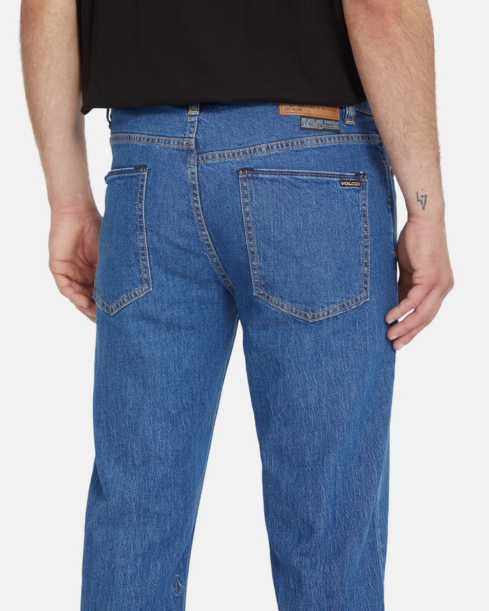 A person is shown from the back wearing Volcom Men's Solver Denim Pants in blue with two back pockets, a brown leather patch on the waistband, and a black shirt. A small tattoo appears on their right arm.