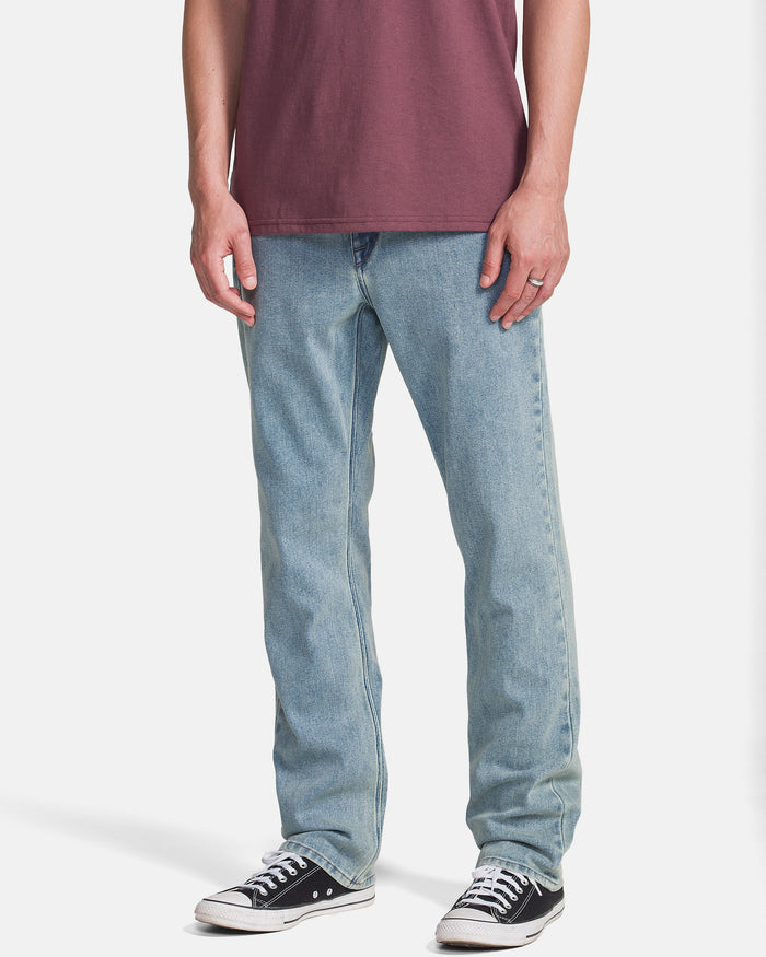 A person wearing Volcom Men's Solver Denim Pants in Dust Bowl Indigo, a maroon t-shirt, and black Converse sneakers stands against a white background. The photo is from shoulders down with arms relaxed at their sides.