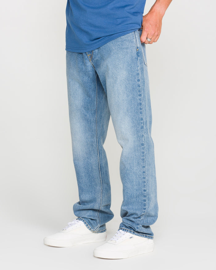A person wearing Volcom Men's Solver Denim Pants in Freedom Blue, paired with a blue shirt and white sneakers, is standing against a plain light background. Only the lower half of the body is visible.
