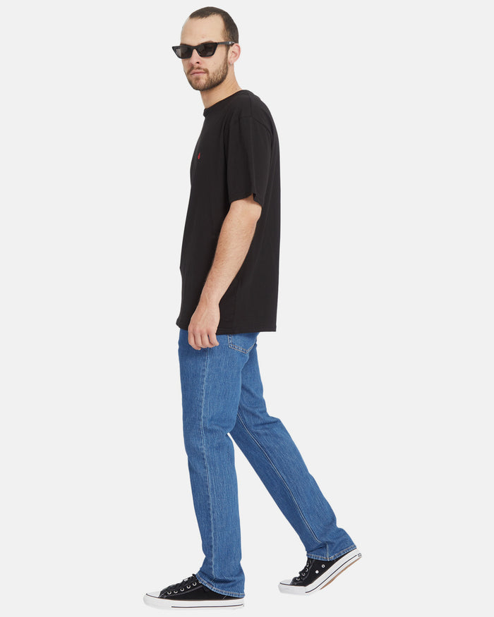 A man in sunglasses, a loose black t-shirt, Volcom Men's Solver Denim Pants, and black sneakers stands against a plain white background, glancing over his shoulder. His sustainable denim blends style with eco-friendly innovation.