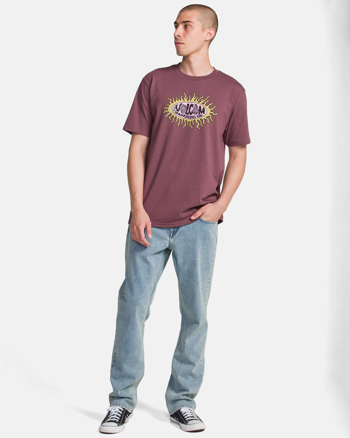 A person wearing a mauve graphic t-shirt, Volcom Men's Solver Denim Pants in light blue, and black sneakers stands with one hand in their pocket, looking to the side against a plain white background.