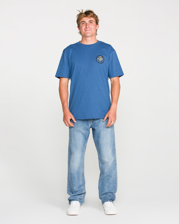 A person stands facing forward, wearing a blue T-shirt with a small circular graphic on the left chest, Volcom Men's Solver Denim Pants in light blue stretch denim with a modern straight leg fit, and white sneakers, against a plain light background.
