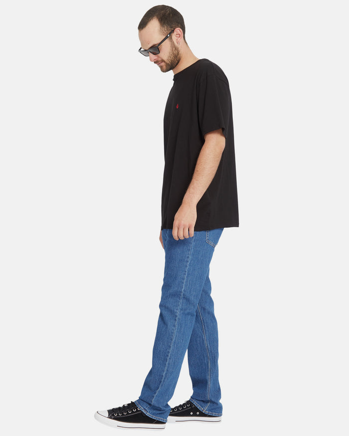 A man in sunglasses, a black t-shirt, Volcom Men's Solver Denim Pants, and black sneakers stands in profile against a plain white background.