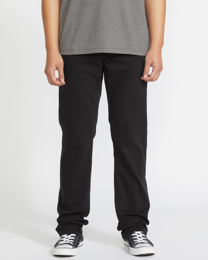 A person is seen from the waist down, wearing a gray t-shirt, Volcom Men's Solver Denim Pant in black, and black-and-white sneakers against a plain white background.