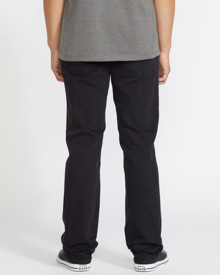 A person faces away from the camera, wearing a gray t-shirt, Volcom Men's Solver Denim Pant in black, and black-and-white sneakers, posed against a plain white background.
