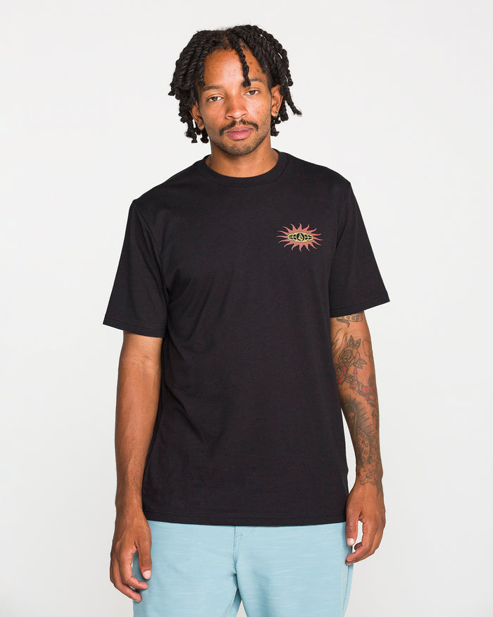 A person with short dreadlocks wears the Volcom Men's Spf91 Short Sleeve Tee, a black, medium-weight heather t-shirt with a small colorful sun graphic on the chest, paired with light blue shorts and standing against a plain white background. This modern fit tee is crafted from a soft ringspun cotton-poly blend, delivering both comfort and style.