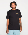 A person with short dreadlocks wears the Volcom Men's Spf91 Short Sleeve Tee, a black, medium-weight heather t-shirt with a small colorful sun graphic on the chest, paired with light blue shorts and standing against a plain white background. This modern fit tee is crafted from a soft ringspun cotton-poly blend, delivering both comfort and style.