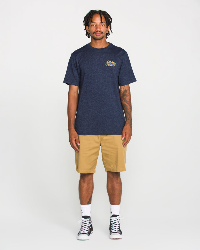 A person stands facing forward, wearing a Volcom Men's Spf91 Short Sleeve Tee in navy with a sun graphic on the chest, paired with khaki shorts, white socks, and black high-top sneakers. The background is plain white.
