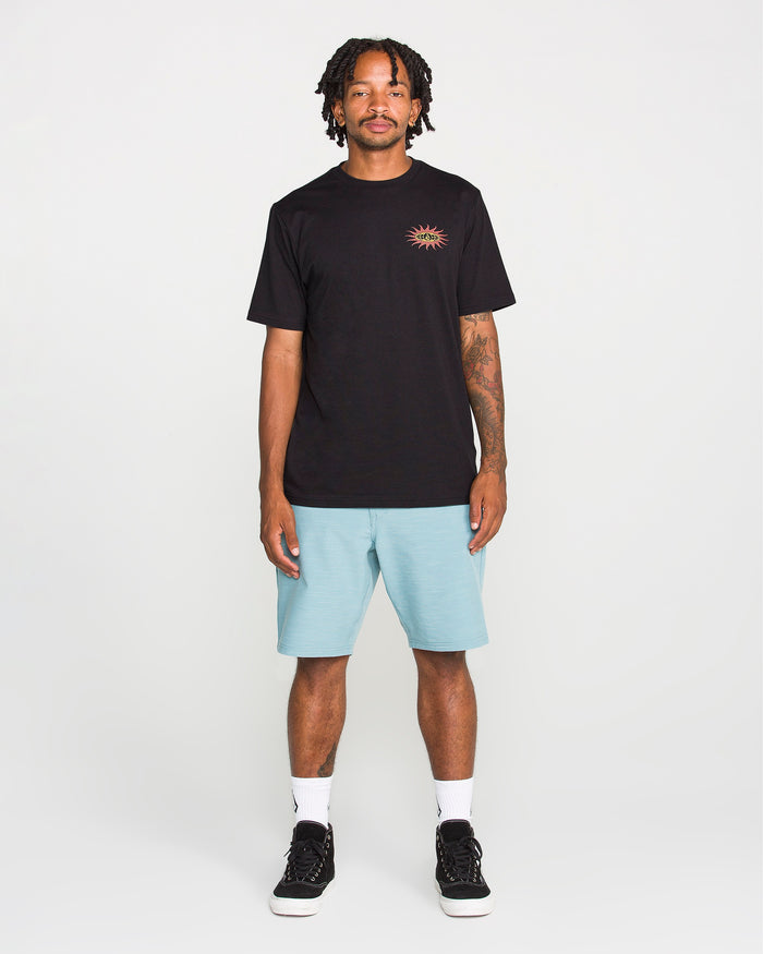 A man stands facing forward against a plain white background, wearing the Volcom Men's Spf91 Short Sleeve Tee—a medium-weight heather shirt with a small sun graphic—paired with light blue shorts, white socks, and black sneakers. The tee features a modern fit and is crafted from a soft ring spun cotton-poly blend for everyday comfort.