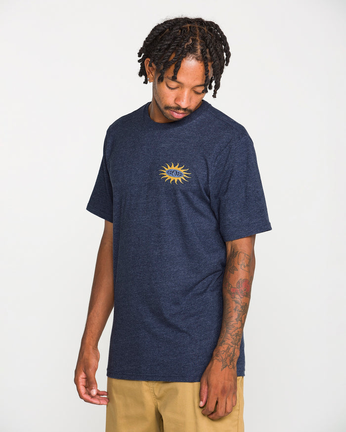 A man stands against a plain white background, looking down with his right hand by his side. He wears tan pants and the Volcom Men's Spf91 Short Sleeve Tee—a navy blue, medium-weight heather shirt featuring a yellow sun graphic on the chest. His left arm is adorned with tattoos, completing the perfect laid-back look in this classic tee.