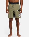 A person stands barefoot against a white background, wearing Volcom Men's St4R G4Zer Mod 20 Trunks in olive green with a black waistband and bold white geometric logos, reflecting the brand’s relaxed colorway style.