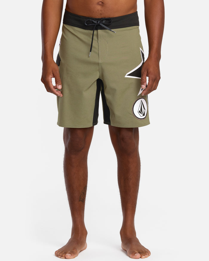 A person stands barefoot against a white background, wearing Volcom Men's St4R G4Zer Mod 20 Trunks in olive green with a black waistband and bold white geometric logos, reflecting the brand’s relaxed colorway style.