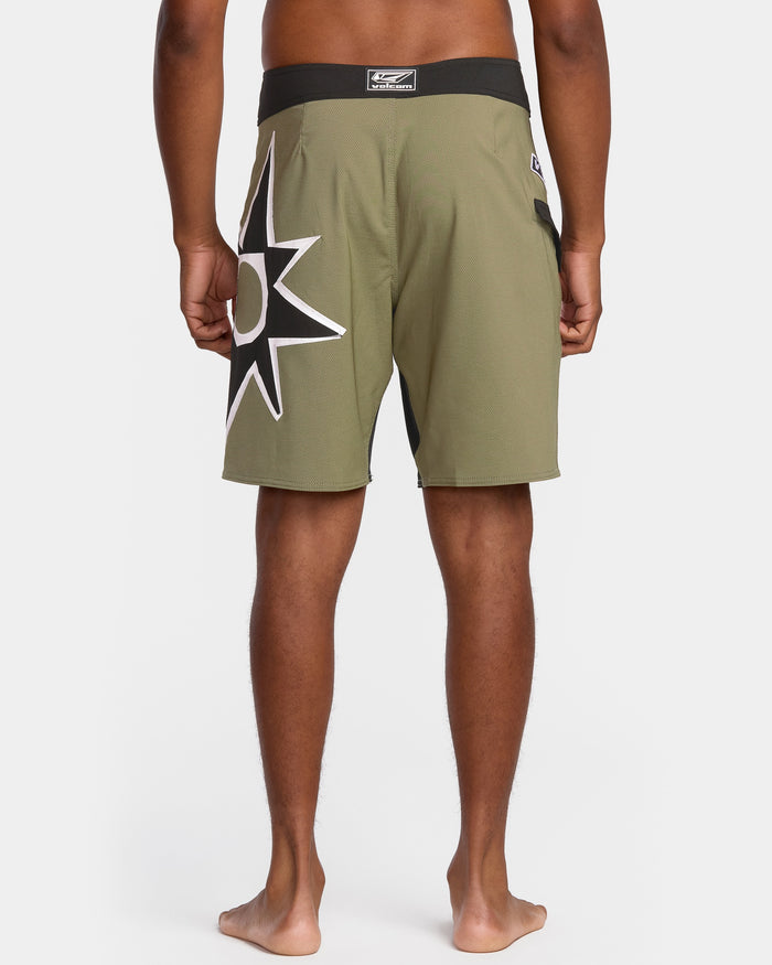 A person stands barefoot, facing away, wearing Volcom Men's St4R G4Zer Mod 20 Trunks in olive green with a bold black and white starburst on the left leg, featuring a black waistband and small logo label at the center back.