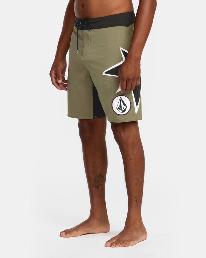 A person stands barefoot in Volcom Men's St4R G4Zer Mod 20 Trunks—olive green with a black waistband, large white star, and circular logo on the left leg—showcasing this sustainable style against a plain white background.