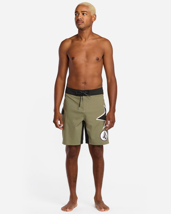 A shirtless, barefoot man with light blonde hair models Volcom Men's St4R G4Zer Mod 20 Trunks in olive green and black with a white geometric design, set against a plain white background.
