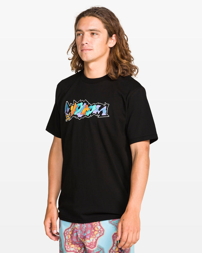 A person with long brown hair wears the Volcom Men's Star Shields Featured Artist Exp Short Sleeve Tee, a black cotton shirt with a colorful graphic, and patterned blue shorts, standing against a white background. This tee adds bold style to the look.