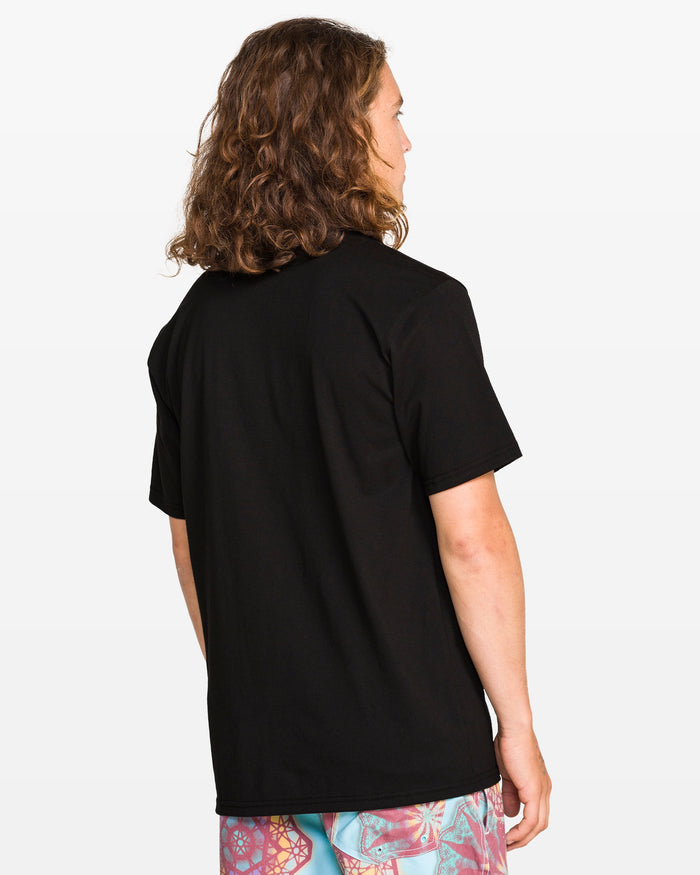 A person with long curly hair stands with their back to the camera, wearing the Volcom Men's Star Shields Featured Artist Exp Short Sleeve Tee and colorful patterned shorts against a white background.