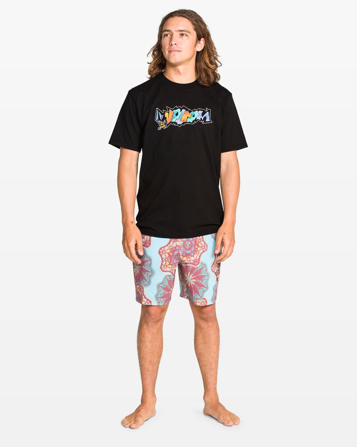 A young man with long hair stands barefoot, wearing a Volcom Men's Star Shields Featured Artist Exp Short Sleeve Tee and patterned pink and blue shorts, set against a plain white background.
