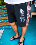 A person wears Volcom Men's Star Shields Featured Artist Mod boardshorts, featuring black fabric with white graffiti-style designs and red patterned side panels, standing on a blue floor with steps and a colorful mat in the background.