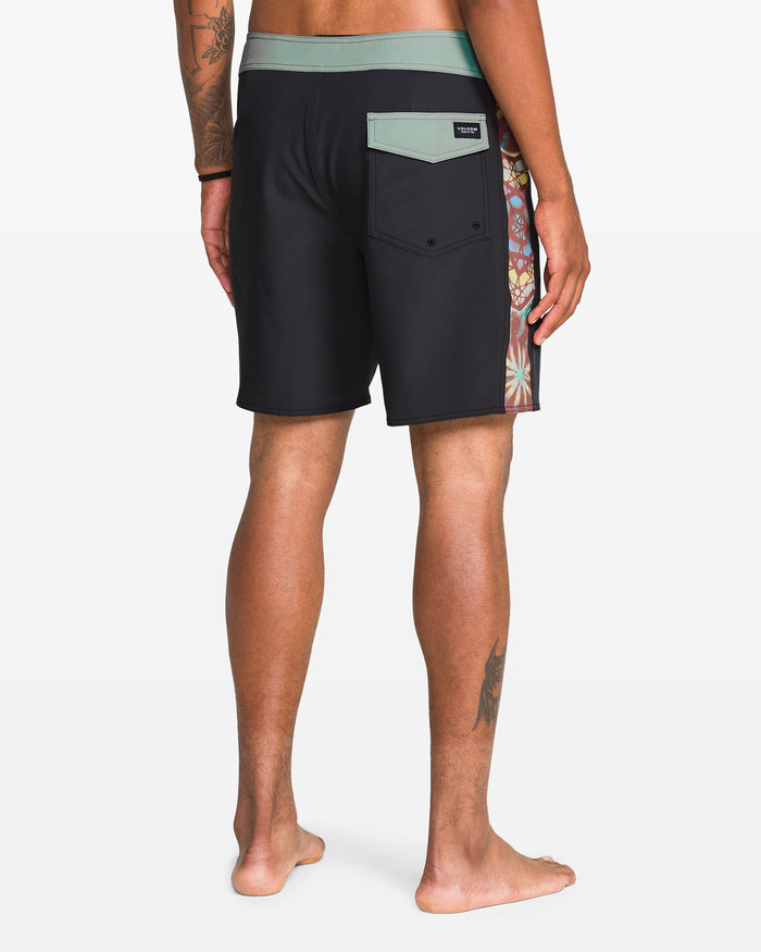 A person with a leg tattoo and medium skin tone stands barefoot, facing away, wearing Volcom Men's Star Shields Featured Artist Mod boardshorts with a green waistband, back flap pocket, and colorful patterned side panel.