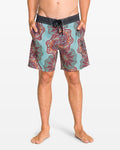 A man stands barefoot with hands in pockets, wearing Volcom Men's Star Shields Featured Artist Pcs Stoney trunks in teal and maroon patterns with a black waistband. This standout style pops against the plain white background.