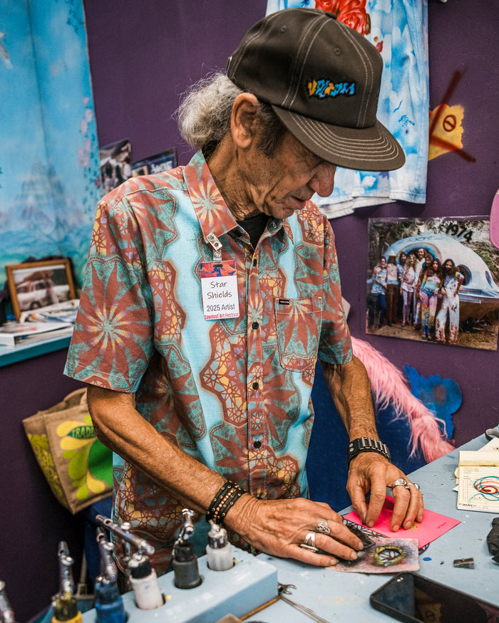 An older man wearing a Volcom Men's Star Shields Featured Artist Short Sleeve Shirt and cap works on an art project at a cluttered table with tools and paints, colorful artwork, and family photos displayed on the wall behind him.