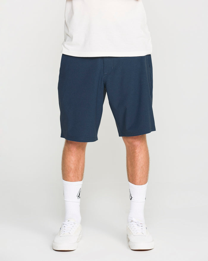 A person wearing a white shirt, Volcom Men's Frickin Cross Shred Static Shorts in navy blue with a 20-inch outseam, white crew socks with a logo, and white sneakers, standing against a plain white background.