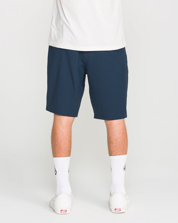 A person stands facing away, wearing a white shirt and Volcom Men's Frickin Cross Shred Static Shorts in navy blue with a 20-inch outseam, paired with white crew socks and white sneakers against a plain background.