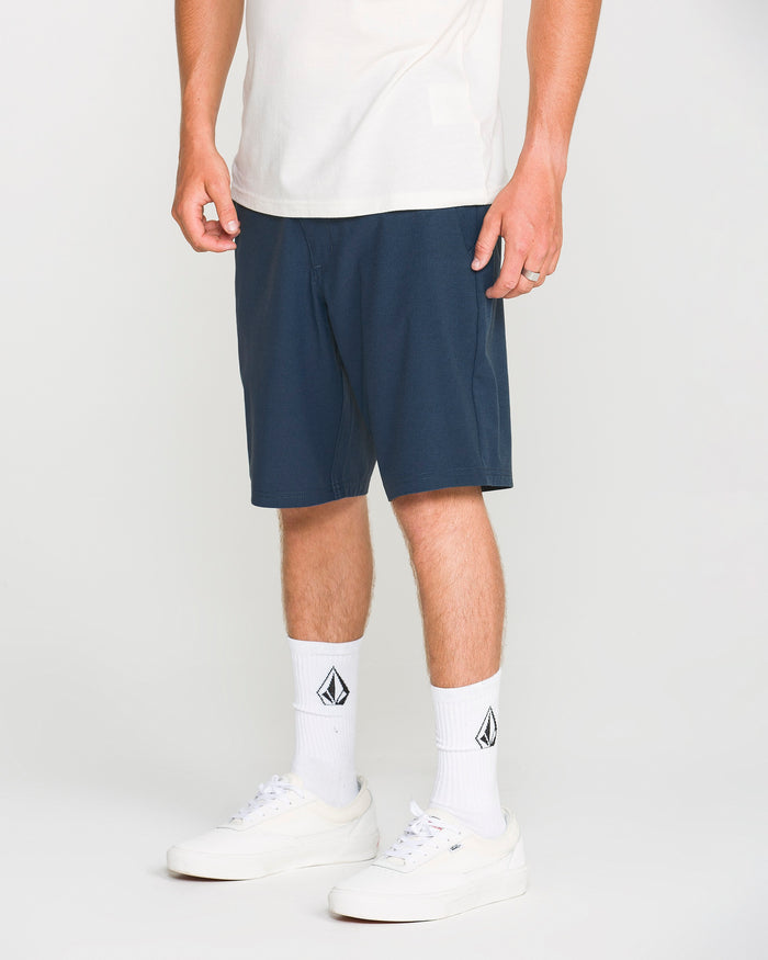 A person stands against a plain background, showing only the lower half of their body, wearing white sneakers, white socks with a black logo, a white t-shirt, and Volcom Men's Frickin Cross Shred Static Shorts in navy featuring a 20-inch outseam.