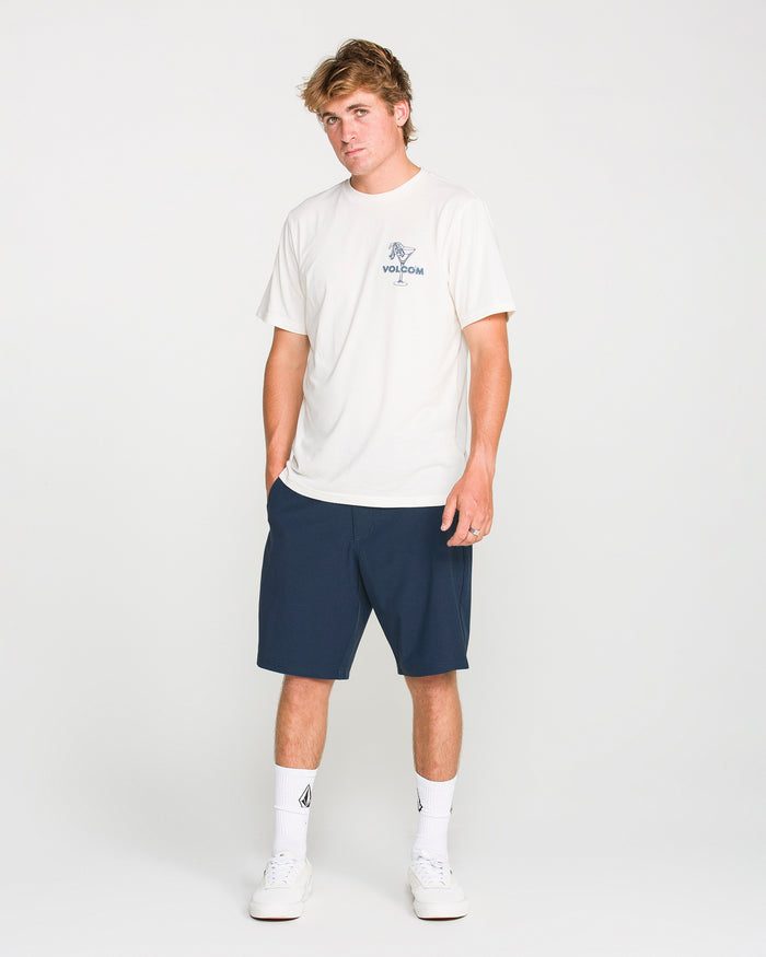 A young man stands facing forward against a plain white background, hands casually in his pockets. He is wearing a white T-shirt paired with Volcom Men's Frickin Cross Shred Static Shorts in navy blue, featuring a 20-inch outseam, and completes the look with white socks and white sneakers.