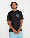 A tattooed man wears the Volcom Men's Still Spiraling Short Sleeve Tee in washed black, which features a blue circular logo on the chest, paired with light blue shorts. The soft hand screen print on the modern-fit heather tee gives a subtle, contemporary touch. He stands against a plain white background, looking at the camera with his hands clasped in front of him.