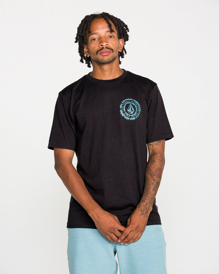A tattooed man wears the Volcom Men's Still Spiraling Short Sleeve Tee in washed black, which features a blue circular logo on the chest, paired with light blue shorts. The soft hand screen print on the modern-fit heather tee gives a subtle, contemporary touch. He stands against a plain white background, looking at the camera with his hands clasped in front of him.