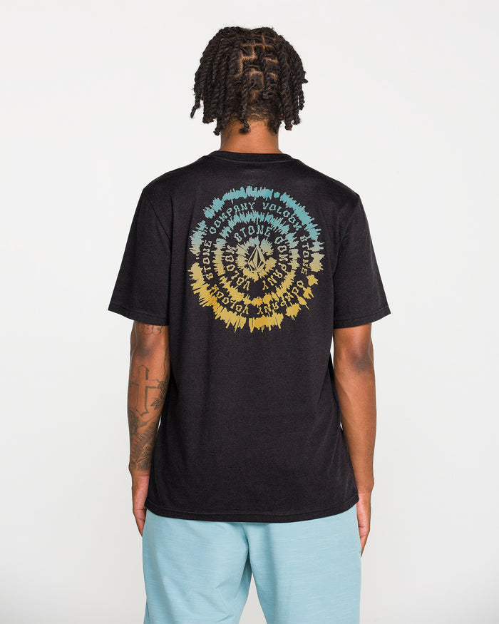 A person with short dreadlocks wears the Volcom Men's Still Spiraling Short Sleeve Tee, a washed black t-shirt featuring a colorful circular graphic and wavy text created with a soft hand screen print on the back. They pair it with light blue shorts while standing against a plain white background.