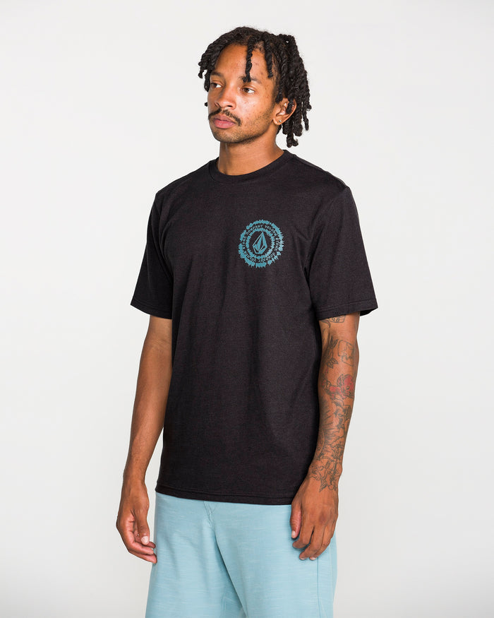 A man with short dreadlocks wears the Volcom Men's Still Spiraling Short Sleeve Tee in washed black, which features a small blue graphic done in a soft hand screen print on the left chest. He pairs it with light blue shorts and stands against a plain white background.