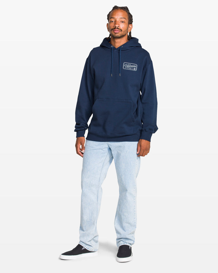 A man with braided hair and a relaxed expression wears the Volcom Men's Stone Coast Pullover in navy, made from brushed fleece and recycled polyester, paired with light blue jeans and black slip-on shoes, against a plain white background.