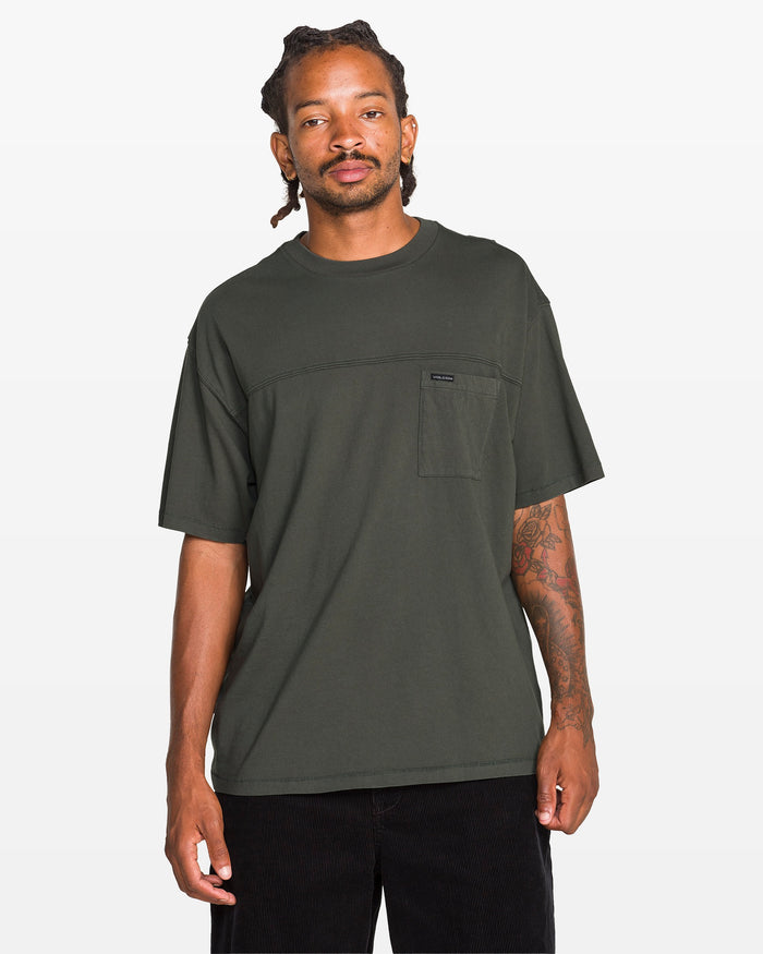 A man with braided hair and tattoos on his left arm wears the Volcom Men's Stone Cruiser Crew t-shirt in dark green, featuring 100% cotton jersey and a small chest pocket, paired with black pants against a white background.