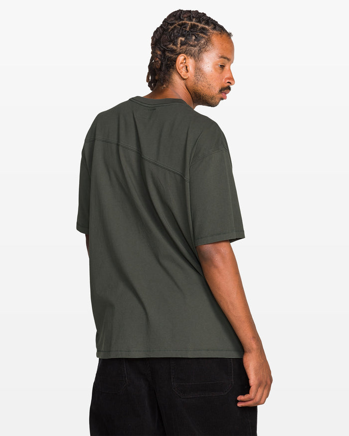 A person with braided hair stands sideways, looking over their shoulder, wearing a loose-fitting Volcom Men's Stone Cruiser Crew T-shirt in 100% cotton jersey (dark green) and black pants against a plain white background.
