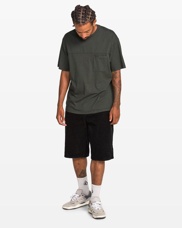 A man faces slightly downward, wearing a dark green Volcom Men's Stone Cruiser Crew tee with a chest pocket, black shorts, white socks, and light grey sneakers against a plain white background.