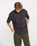 A person with light brown hair wearing the Volcom Men's Stone Optix Polo in black, which features a boxy fit and a yarn-dyed jacquard pattern, paired with olive green pants, looks down while adjusting their shirt against a plain white background.