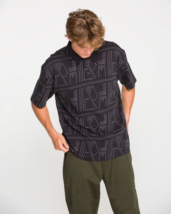 A person with light brown hair wearing the Volcom Men's Stone Optix Polo in black, which features a boxy fit and a yarn-dyed jacquard pattern, paired with olive green pants, looks down while adjusting their shirt against a plain white background.