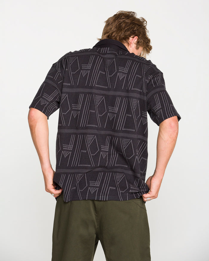 A person with light brown hair faces away, wearing the Volcom Men's Stone Optix Polo—a black patterned yarn-dyed jacquard short-sleeve shirt with a boxy fit—and olive green pants, standing against a plain white background.