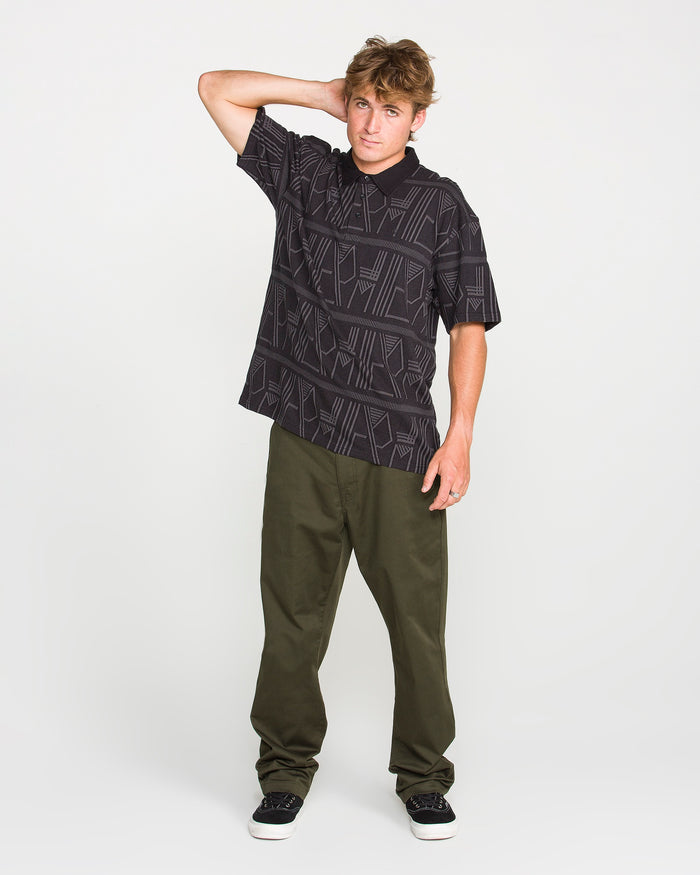 A young man wearing the Volcom Men's Stone Optix Polo—black with a distinctive pattern, boxy fit, and crafted from yarn-dyed jacquard—and olive green pants stands with one hand behind his head and the other by his side, posing against a plain white background.