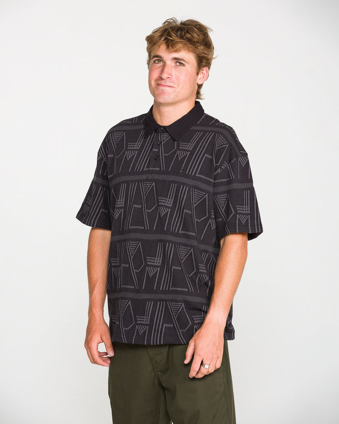 A person with short brown hair wearing the Volcom Men's Stone Optix Polo in a black, boxy fit crafted from yarn-dyed jacquard, paired with olive green pants, stands against a plain white background and looks at the camera with a neutral expression.