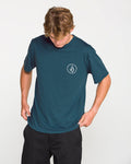 A man wearing the Volcom Men's Stone Tech Short Sleeve Tee in teal, featuring a small white geometric logo on the left chest, looks down as he adjusts the hem. Crafted from quick-dry fabric with a modern fit, the T-shirt is styled with black pants against a plain white background.