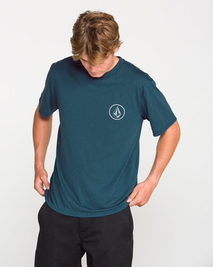 A man wearing the Volcom Men's Stone Tech Short Sleeve Tee in teal, featuring a small white geometric logo on the left chest, looks down as he adjusts the hem. Crafted from quick-dry fabric with a modern fit, the T-shirt is styled with black pants against a plain white background.