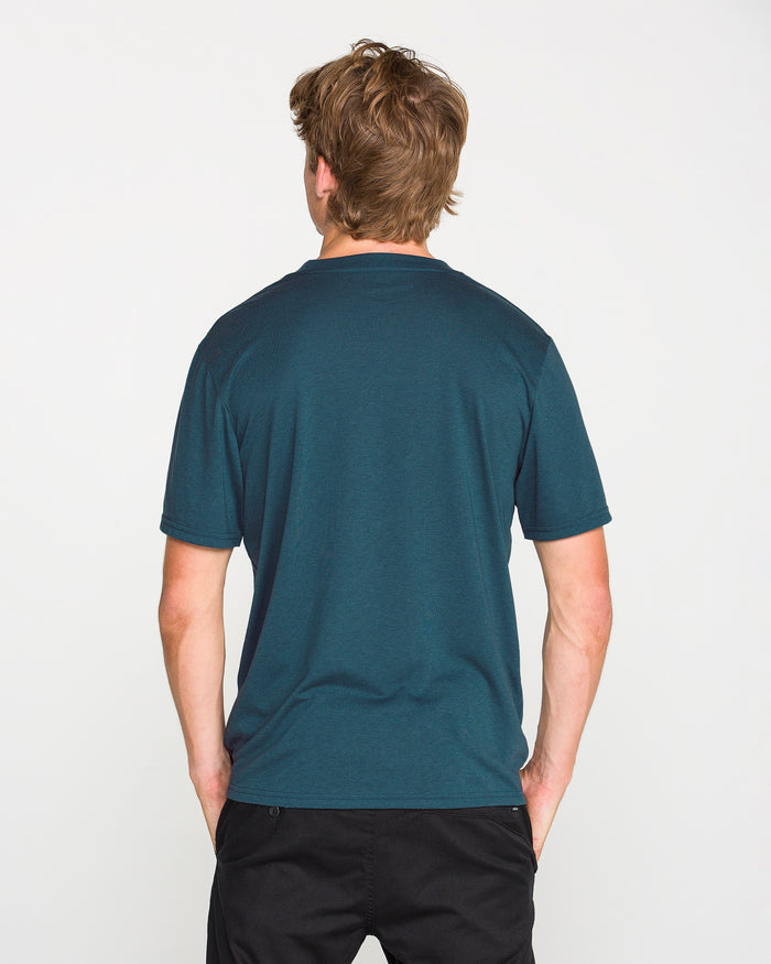 A person with light brown hair is standing with their back to the camera, wearing the Volcom Men's Stone Tech Short Sleeve Tee in teal—crafted from quick-dry fabric—paired with black pants, against a plain white background.