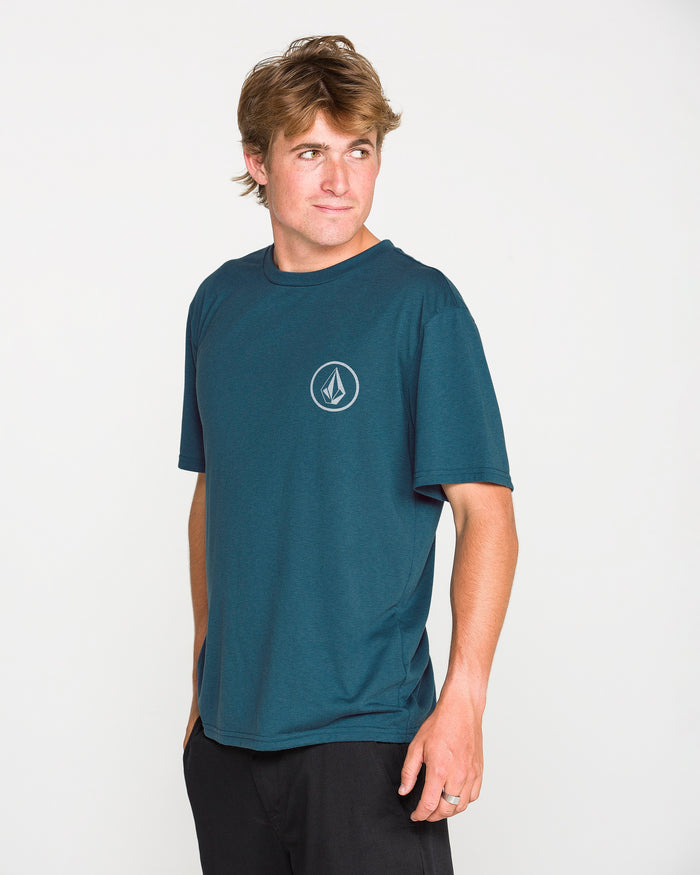 A person with light brown hair wears the Volcom Men's Stone Tech Short Sleeve Tee—a teal short-sleeved shirt with a small chest logo, crafted from quick-dry fabric for all-day comfort and featuring a modern fit—paired with black pants. They stand against a plain white background, looking to the side.