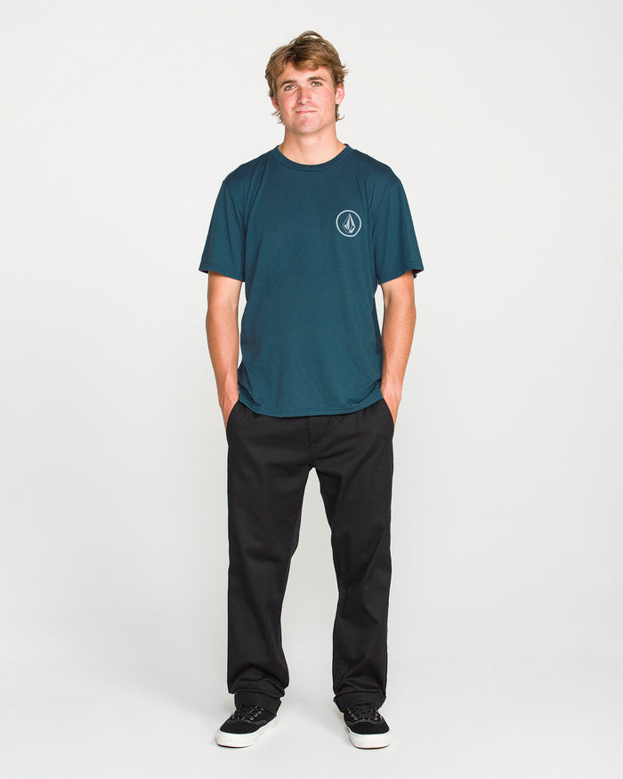 A young man stands facing forward with hands in his pockets, wearing the Volcom Men's Stone Tech Short Sleeve Tee in teal—crafted from quick-dry fabric and featuring a small logo—paired with black pants and black sneakers, against a plain white background.
