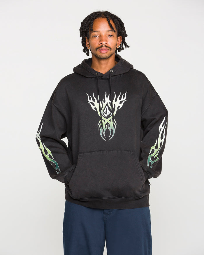 A person wearing the Volcom Men's Stone Trip Pullover in washed black, featuring white and green flame designs on the chest and sleeves, stands against a plain white background.