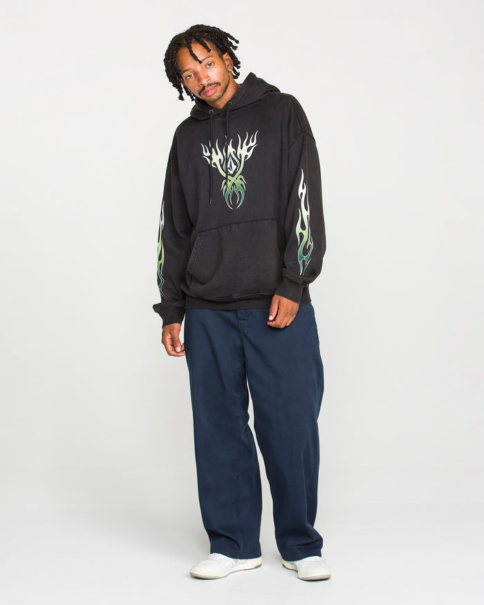 A person stands against a plain background wearing the Volcom Men's Stone Trip Pullover, a washed-black tribal hoodie featuring striking green flame designs. Paired with navy blue pants and white shoes, they pose relaxedly with a slight smile. The heavyweight fleece fabric of the hoodie adds both comfort and style to the overall look.